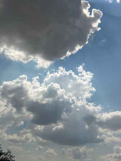 The media file shows a cloudy sky with the sun partially visible, creating dramatic light rays. The clouds vary in density, with some appearing dark and heavy, suggesting potential rain, while others are bright white and puffy, illuminated by the sun. The sky is a clear blue, peeking through the clouds in patches. The overall scene evokes a sense of grandeur and the dynamic nature of weather. The lighting suggests it is daytime, likely late afternoon given the angle of the sun's rays. There are no visible people or objects, and no text. The image appears to be a photograph taken from the ground looking upwards into the sky. The provided location context of Загорци, Bulgaria, indicates a rural or semi-rural setting.