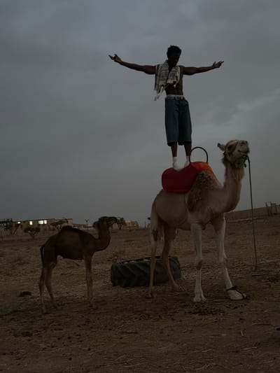 In Douar Ait Daoud, Morocco, a young man stands with outstretched arms atop a camel, his feet balanced on the hump. He is shirtless, wearing blue shorts and a scarf around his neck. The camel, with a red saddle, stands on dusty ground. To the left, a smaller camel calf stands nearby. In the background, buildings and trees are visible under a cloudy, overcast sky. The overall scene suggests a remote, arid environment.