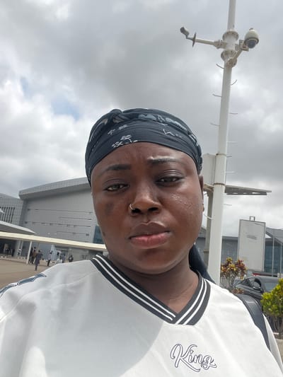A close-up, waist-up portrait captures an individual in an outdoor setting, taken from a low angle and rotated 90 degrees clockwise. The subject is a person of color, looking directly into the camera with a neutral expression. They wear a dark, patterned bandana covering their head and a white top featuring black and white stripes around the collar. The word "Kings" is visibly embroidered in a stylized font on the left side of their chest (viewer's right). A small silver nose ring adorns their right nostril.
The background is dominated by an overcast sky filled with grey clouds. A prominent white pole, fitted with a surveillance camera or weather monitoring equipment, stands to the left of the subject. This equipment includes a spherical camera component and several arm-like extensions. In the mid-ground, modern buildings with glass exteriors are visible, suggesting an urban or commercial area. A dark vehicle and some green foliage are partially seen further back on the right. Distant, blurry figures can be discerned near one of the buildings. The photograph depicts a daytime scene under cloudy weather conditions in Gwaska, Nigeria.