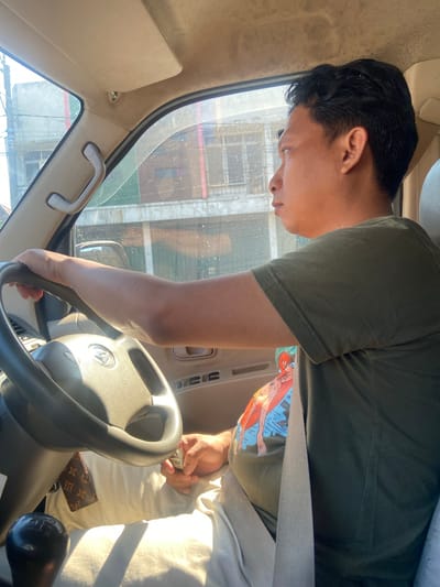 A man is driving a vehicle in Ngrame, Indonesia. He is wearing a green t-shirt and has dark hair. The man is holding the steering wheel with his left hand, and his right hand is resting on his lap. The car's interior is visible, including the dashboard, steering wheel, gear shift, and seatbelt. Sunlight is streaming in through the windows, casting shadows on the interior. Outside the car, buildings and trees are visible. The man appears to be looking straight ahead, focused on the road.