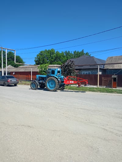 In the city of Avtury, Russia, on a clear sunny day, a blue tractor with a red mowing attachment is parked on the side of a paved road. The tractor, which appears to be old and industrial, has large, rugged tires. The mowing attachment, featuring two circular blades, is positioned behind the tractor. To the left of the tractor, a dark gray car is parked. In the background, there are residential buildings with various roofing materials, including corrugated metal and gray shingles. Trees with green foliage line the property behind the buildings, and electrical wires are strung overhead. The road surface is primarily asphalt with visible stones and texture.