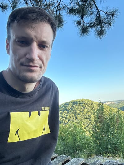 A man stands in front of a wooded hillside under a clear blue sky, with pine branches overhead. He is wearing a dark t-shirt with a yellow graphic and the words "THE ORIGINAL" printed above it. In the background, a densely forested mountain landscape stretches out, with some buildings visible in the distance. The scene appears to be captured from a slightly elevated position, possibly a viewpoint or a park in Pryvorottia Druhe, Ukraine.