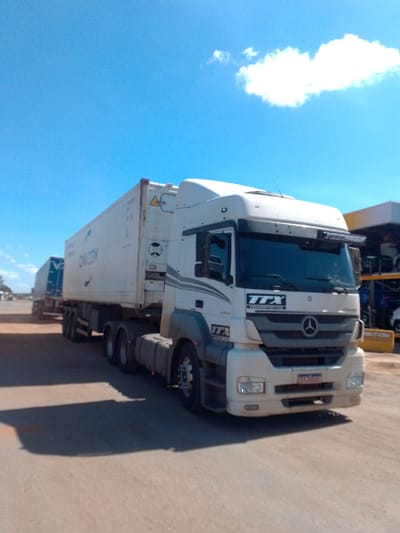 A white Mercedes-Benz semi-truck with a white shipping container is parked on a dirt road. The truck has "TTX TRANSPORTES" written on its side. In the background, another truck with a blue shipping container is visible, along with buildings and trees. The sky is clear and blue with a single cloud. The image appears to have been taken in Cáceres, Brazil.