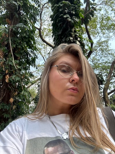 A person with blonde hair and round glasses gazes towards the camera. The individual wears a white t-shirt and a silver necklace with a cross-shaped pendant. Lush greenery, including tree branches and foliage, surrounds the subject. The background suggests an outdoor setting, possibly a park or garden. The photo's angle is slightly upward, capturing the person's face and upper body.