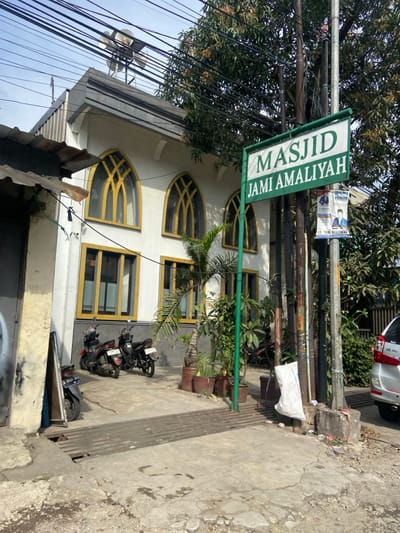 A green sign with white lettering reads "MASJID JAMI AMALIYAH" in front of a mosque in Kecamatan Dayeuhkolot, Indonesia. The mosque is a white, single-story building with four arched windows featuring yellow frames. There are three motorcycles parked in front of the mosque and potted plants. The street in front of the mosque is paved with gray concrete and lined with a row of brown wooden poles. The image is captured from a street level perspective, looking toward the mosque.