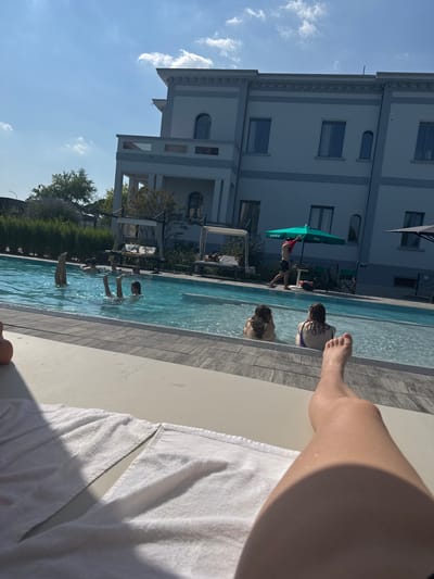 The image depicts a bright, sunny day at an outdoor swimming pool. The perspective is from someone lying on a lounge chair, facing towards the pool. The scene is populated with several people enjoying the water. In the foreground, a person's legs and feet are visible, along with a white towel draped over a lounge chair.
Further into the pool, two individuals are seen, one partially submerged and the other with her head above water. In the background, there are more people in the pool and on the poolside. One person is standing near a green umbrella, wearing a red shirt and dark shorts. A white building with multiple windows and arched details stands behind the pool area, suggesting a hotel or resort. Lush green hedges and trees line the edge of the pool, adding to the serene atmosphere. The clear blue sky with scattered white clouds indicates pleasant weather, likely midday given the strong shadows. The overall mood appears relaxed and recreational, with people enjoying a summer day by the pool. No visible text is present in the image.