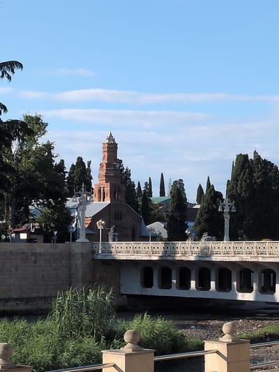 The image depicts a scene in Ganja City, Azerbaijan, featuring a distinctive brick church with a tall, octagonal tower topped with a spire. The tower has a clock face visible. The church has a pitched roof and arched windows.
In the foreground, a modern bridge with a decorative railing spans a body of water. The bridge's underside is supported by multiple arched structures. Along the bridge's railing, lampposts are visible.
To the left of the church, a stone wall forms the embankment of the river or canal. Lush green vegetation, including tall grass and trees, surrounds the area. The sky is blue with scattered white clouds. The overall impression is a sunny day in a city with historical and modern architectural elements.