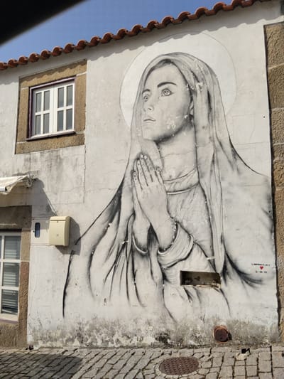The image shows a large black and white mural on the side of a building. The mural depicts a woman with her hands clasped in prayer, wearing a head covering and a halo around her head. The building is made of light-colored stone and has a tiled roof. There is a window to the left of the mural. Below the mural there is an empty rectangular opening in the wall and an "AGUA" marked manhole cover on the street. A signature and date "L.EMPREINTE.JO.V 15-08-23" are written in red at the lower right of the mural. The background sky is blue. The location is São João da Pesqueira, Portugal.