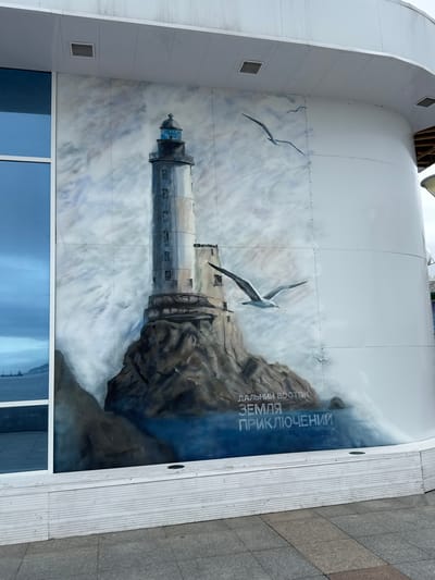 The image displays a mural painted on the exterior curved wall of a building, situated in the city of Russky, Russia. The mural depicts a lighthouse situated on a rocky outcrop, with waves crashing around its base. Two seagulls are painted in flight near the lighthouse. The background consists of a cloudy sky rendered in shades of blue and white. Below the mural, text is visible in Cyrillic letters. To the left of the mural is a large, reflective blue window. The building appears to be modern, with a white facade. The ground in front of the building is paved with grey and brown tiles. The scene is captured from a slightly elevated perspective, looking up at the mural.