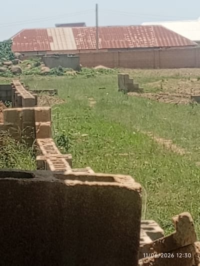 The image depicts an outdoor scene in DU, Nigeria, likely a construction site. In the foreground, partially visible brickwork suggests the beginnings of a structure, with a large, dark, textured object dominating the bottom left. Behind this, a row of partially constructed brick walls extends into the middle ground, with visible brick and mortar. Further back, a concrete step-like structure stands in a grassy area. The background features a long, brick wall, and beyond that, a building with a corrugated metal roof, showing signs of age and rust. The overall environment is characterized by dry grass, scattered debris, and a clear sky. The timestamp "11/06/2026 12:30" is visible in the bottom right corner. There are no people or animals visible in the image.