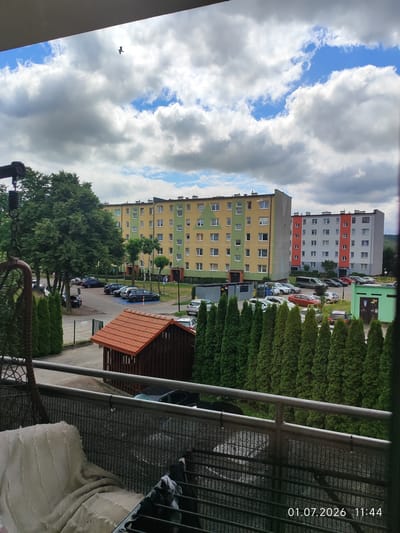 The image shows a view from a balcony overlooking a residential area in Lębork, Poland, with apartment buildings, parked cars, trees, and a small wooden shed. The sky is partially cloudy with blue patches. The timestamp "01.07.2026 11:44" is visible in the bottom right corner. In the foreground, a white blanket is draped over a railing, and a black mesh fence is also visible. In the middle ground, a small wooden shed with a terracotta roof is situated next to a row of tall, dark green coniferous trees. Beyond the trees, a parking lot is filled with various cars, and several multi-story apartment buildings are visible. The apartment buildings are painted in different colors, including yellow, green, orange, and gray. A single bird is seen flying in the sky.