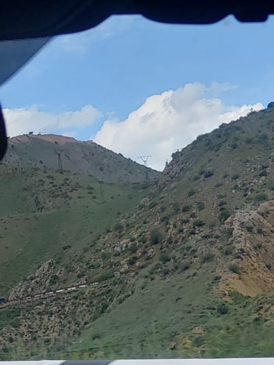 A vibrant landscape shot captured from what appears to be a moving vehicle depicts a mountainous region under a bright blue sky. A dark, out-of-focus element, likely part of a car's interior or window frame, occupies the left edge and bottom-left corner of the image, framing the view.

The central focus is a large, undulating mountain range with slopes covered in a mixture of lush green grass and rocky outcrops. Patches of lighter, bare earth are also visible on some of the slopes. The sky above is a brilliant azure, dotted with fluffy, white cumulus clouds.

Several man-made structures are integrated into the natural environment. On the higher, more distant ridges, slender, metallic lattice towers, possibly electricity pylons or cable supports, punctuate the skyline. Further down on the right-hand slope, a long, thin, linear structure, resembling a narrow track or a section of pipeline, stretches horizontally across the terrain.

The scene is well-lit, indicating a clear, sunny day, likely in the late morning or early afternoon. The overall impression is one of vastness and natural beauty, characteristic of mountainous regions. The presence of infrastructure suggests human activity within this rugged landscape. The location is identified as Tigranashen, Armenia. There is no discernible text in the image.