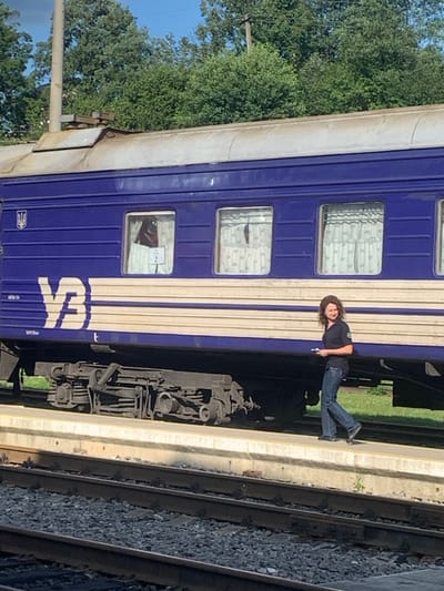 The scene depicts a train station in Yasinia, Ukraine, under clear daylight conditions. A dark blue and beige passenger train car, bearing the white "УЗ" (Ukrzaliznytsia) logo and a Ukrainian trident emblem, occupies the upper half of the frame. Three windows are visible on the train car, each fitted with white, patterned curtains. The leftmost window displays a dark, possibly reddish object behind the curtain and a white paper sign marked with the number "2." The train's metal bogie is visible beneath the car.

On the concrete train platform adjacent to the train, an individual with shoulder-length curly brown hair is walking from left to right. The person is wearing a dark, short-sleeved collared shirt with a circular emblem on the left chest and blue jeans. A small, light-colored object is held in their left hand. The individual's gaze is directed towards the right side of the frame.

Below the platform, multiple railway tracks are visible, consisting of metal rails laid on gravel ballast. In the background, beyond the train and platform, dense green foliage and a clear blue sky are present. A tall, dark pole is partially visible on the far left, extending upwards.