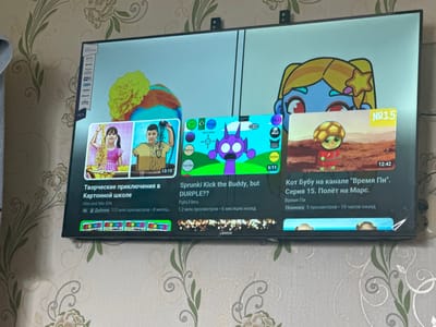The image shows a television screen displaying various YouTube videos. The TV is mounted on a wall with floral wallpaper. On the left side of the screen, a video titled "Творческие приключения в Картонной школе" (Creative Adventures in Cardboard School) is featured, with a thumbnail showing two children. Below this, the video has 4K resolution and 112 million views, posted 8 months ago by "Vlad and Niki IDN".
In the center of the screen, a video titled "Sprunki Kick the Buddy, but DURPLE??" is displayed. Its thumbnail shows a purple cartoon character with big eyes. This video has 1.2 million views and was posted 6 months ago by "PghlFilms".
On the right side, a video titled "Кот Бубу на канале 'Время Пи'. Серия 15. Полёт на Марс." (Cat Booboo on the "Time Pi" channel. Episode 15. Flight to Mars.) is shown. The thumbnail features a cartoon turtle character. This video is a new release with 5 views and was posted 19 hours ago.
A sidebar on the far left of the TV displays app icons including Netflix, YouTube, Prime Video, and Wi-Fi. The television is a smart TV with "webOS Hub" indicated at the top. The bottom of the television has a "LENOX" logo. The overall scene suggests a living room environment.