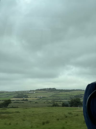 This media file captures a view from a moving vehicle, likely a train, looking out at a landscape in Carrowcannon Lower, Ireland. The perspective is from inside the vehicle, with the top right corner of the frame showing a dark blue hat or headwear.
The scene is dominated by a vast expanse of overcast sky, which takes up the majority of the left side of the image. The sky is a uniform light grey, suggesting a cloudy or rainy day. On the right side, a rolling green landscape unfolds. The land is divided into various fields, demarcated by dark lines that could be hedges or stone walls. Patches of trees and some white buildings, possibly houses or farm structures, are scattered across the hillside, indicating a rural setting. The overall impression is one of lush greenery, characteristic of the Irish countryside.
The time of day appears to be daytime, given the natural light, but the overcast conditions make it difficult to determine the exact hour. The weather is clearly not sunny, and the mood evoked is somewhat subdued due to the dull sky. The activity taking place is incidental to the landscape itself; the viewer is passively observing the scenery as they travel. There is no discernible text within the image.