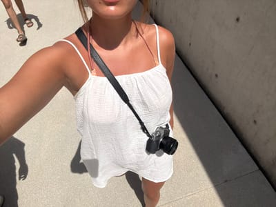 The image depicts an outdoor scene in San Isidro Llano Grande, Mexico, captured from a high-angle perspective. The primary subject, a person with tanned skin, is visible from the upper chest downwards, wearing a white, textured spaghetti-strap garment. A black camera strap diagonally crosses their torso, from which a black digital camera with a lens is suspended at their right hip. The subject's lower lip and chin are visible at the top edge of the frame. The ground is a uniform, light-colored paved surface, consistent with concrete. Strong sunlight casts a distinct shadow of the primary subject onto the ground. In the upper left background, the lower legs and feet of a second individual, wearing brown sandals, are visible walking away, also casting elongated shadows. To the right, a dark gray concrete wall, featuring circular indentations, extends diagonally across the frame, with a significant portion cast in shadow.