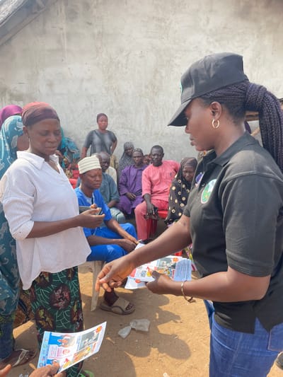 An outdoor scene in Osara Gada, Nigeria, depicts an outreach event or community gathering. In the foreground, a woman wearing a black polo shirt, blue jeans, and a black baseball cap with a green logo is distributing informational flyers. Her right hand extends a stack of colorful leaflets towards a woman on the left, who is wearing a white striped V-neck shirt and a brown headscarf. This woman holds a mobile phone in her left hand and a single flyer in her right. Both women wear earrings; the distributor has a gold bracelet. The ground is exposed dirt, with a crumpled piece of white debris visible. In the midground and background, a group of approximately fifteen individuals, primarily adults, are seated on chairs and benches against a plain, light grey concrete wall. Their attire includes traditional Nigerian garments like agbadas in blue and pink, various head coverings, and casual clothing. They appear to be observing the interaction in the foreground.
