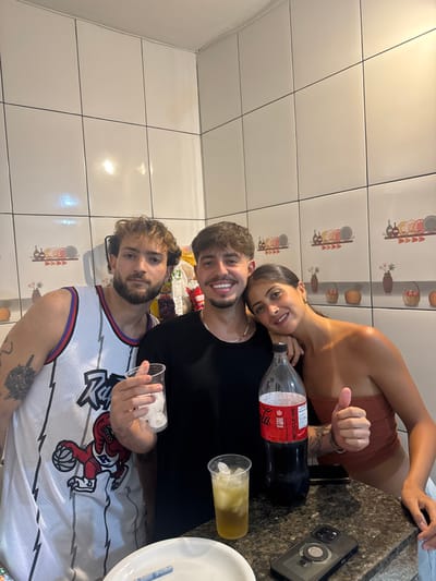 Three young adults pose closely together. One wears a white basketball jersey with a cartoon character on it. Another is wearing a black t-shirt and has a woman leaning onto their shoulder. She wears a brown top and gives a thumbs up. A bottle of Coca-Cola, two plastic cups, a cell phone, and a plate are on the counter. The background is a tiled wall.