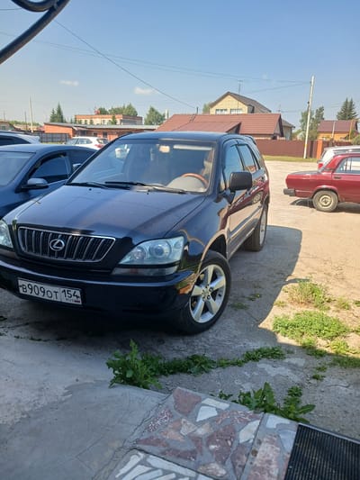 In the city of Prokudskoye, Russia, on a sunny day, a dark blue Lexus RX SUV is parked on a dirt and concrete lot. The SUV is the primary subject, facing slightly towards the viewer and to the left. Its license plate reads "B909OT 154 RUS". To the right of the Lexus, a red sedan is partially visible, also parked. The background consists of several residential buildings with tiled roofs, fences, and trees. Power lines are visible overhead against a clear blue sky. The ground is a mixture of loose dirt, gravel, and cracked concrete, with patches of green vegetation growing through the cracks. The scene appears to be a residential area or an informal parking lot. There are no people visible in the image.