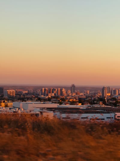 This is an outdoor, vertical, medium shot taken during golden hour, looking towards the city of Be'er-Sheva, Israel. The foreground is dominated by a blur of dry, golden-brown vegetation. Behind this, a line of modern buildings with white facades and dark rectangular windows stretches across the frame. Further in the distance, a cityscape of taller buildings, including high-rises, is visible. The sky transitions from a light blue-grey at the top to a warm orange-yellow gradient as it approaches the horizon, suggesting the sun is low in the sky. The overall impression is of a city seen from a slightly elevated vantage point at sunset.