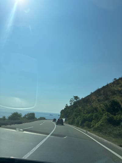 The image depicts a highway in Kanjoši, Montenegro, likely taken from a vehicle moving along the road. The scene shows a winding asphalt road with white lane markings, bordered by a metal guardrail on the right. To the left of the road, a steep, tree-covered hillside rises, with sparse vegetation and rocky patches visible. In the distance, the sea or a large body of water is visible, with a hazy horizon. Several cars are traveling on the highway in the same direction as the camera. The sky is a clear, bright blue, suggesting a sunny day. The sun is visible in the upper left corner of the image, casting a bright glare and lens flare that obscures part of the sky and the hillside. The overall impression is of a scenic drive along a coastal highway. No text is visible in the image.