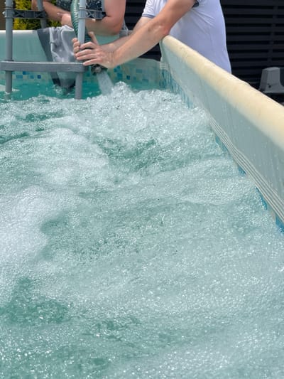Two individuals are in the process of filling a small, inflatable swimming pool with water from a garden hose. The pool is situated outdoors, as indicated by the presence of greenery and a wooden fence in the background. The water in the pool is churning and bubbly, suggesting it is being actively filled. One person's hands are visible, holding the hose as it dispenses water. The other person is partially visible, also near the pool's edge. The water is a clear, turquoise color, and the pool has a tiled pattern on its interior walls. The overall scene depicts a common summer activity. The specific location in Koroviia, Ukraine, cannot be determined from the visual information alone.
