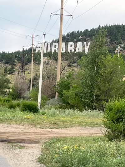 In the city of Shchuchinsk, Kazakhstan, large white letters spelling out "SHCHUCHINSK" are prominently displayed on the side of a hill. The letters are positioned above a line of trees and vegetation that covers the hillside. Power lines and utility poles are visible in the foreground, running across the scene and intersecting with the view of the letters. The landscape appears somewhat arid, with patches of dry grass and scrubby bushes interspersed with areas of greener foliage. A dirt road or path is visible in the lower portion of the image, leading into the frame from the bottom left. The sky is overcast, suggesting a cloudy day. The overall scene depicts a prominent landmark of the city, set against a natural, hilly backdrop with evidence of infrastructure.