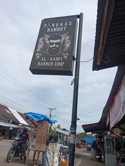 The image captures a street scene in Pulorayeuk, Indonesia. A prominent barber shop sign, "PANGKAS RAMBUT AL-KAHFI BARBER SHOP," hangs from a pole, featuring a graphic of a man with a beard and scissors. To the right, a corrugated metal roof structure extends over a shop. In the foreground, a man wearing a dark purple t-shirt, camouflage shorts, and a hat sits on a motorcycle. Several cardboard boxes are stacked on a wooden stand covered by a blue tarp, suggesting a market stall. Further down the street, more buildings and market activity are visible, with people and vehicles present. The sky is overcast.