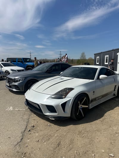 The image depicts a row of parked cars, including a white Nissan 350Z with a "SPORT" graphic, a grey Mercedes-Benz C-Class, and a blue Jeep Wrangler. People are standing in the background near power lines. The cars are parked on a concrete and dirt surface. A building is visible in the background. The location is Sadovoe, Russia.