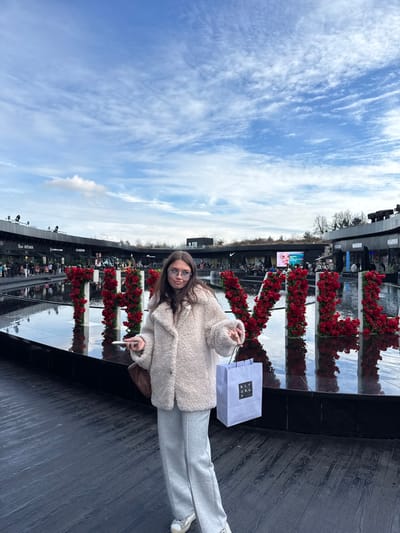 A young adult female is depicted standing in an outdoor commercial setting, identified as The Village outlet shopping center in Villefontaine, France. She has long, dark brown hair and wears light-colored eyeglasses. Her attire consists of a cream-colored, textured fleece jacket, light grey wide-leg trousers, and white sneakers with dark detailing. In her left hand, she holds a white smartphone, and in her right hand, she carries a light blue shopping bag labeled "RITUALS...". A brown shoulder bag is visible on her left hip. She is positioned in front of a large, dark, reflective water feature containing three-dimensional letters constructed from red roses, spelling "THE VILLAGE". The letters "THE VILL" are clearly visible, with the rest extending beyond the right frame edge. The background features several modern, dark-colored commercial buildings with flat, landscaped roofs, housing various retail establishments, including partial signage for "ADIDAS," "NIKE," and "HARIBO." Indistinct pedestrian figures are visible throughout the background. Bare trees stand behind the buildings, suggesting a cold season. The sky is partly cloudy with areas of visible blue. The ground in the foreground is dark-colored plank flooring.