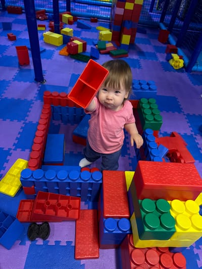 A high-angle shot depicts a toddler playing with oversized building blocks in an indoor playspace. The space features a purple and blue jigsaw-patterned floor. The child, wearing a pink shirt and dark pants, is surrounded by large plastic blocks in red, blue, green, and yellow. The child is holding a red block. Other blocks are arranged to form a low structure around the child. The background shows additional play structures, including netting and a padded climbing area. The location is not specified, but the instruction includes “Перовское сельское поселение, Ukraine,” which is the location.