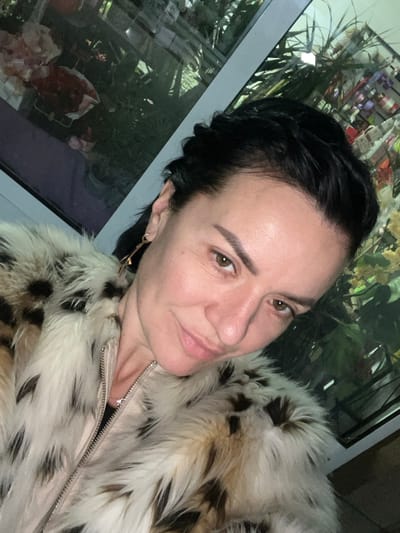 A woman is depicted in a close-up, angled self-portrait, facing slightly towards the viewer with a subtle smile. She has dark hair, styled partially pulled back with some braiding visible on her left side (viewer's right), and brown eyes. Her skin tone is fair. She is wearing a faux fur coat with a cream or off-white base color and dark brown or black leopard-like spots, secured with a visible metallic zipper down the front. A delicate silver-colored chain necklace with a small pendant is visible at her throat. A gold-colored, elongated dangling earring is visible on her left ear (viewer's right). The background consists of a large glass window, showing reflections of what appears to be an interior space. The reflections include green foliage, possibly potted plants or floral arrangements, and various colorful items that resemble merchandise or decorative elements, specifically pink, red, purple, and yellow tones, suggesting a commercial establishment, possibly a florist or gift shop. The window frame is light-colored. The specific location within the city of Smederevska Palanka Municipality, Serbia, cannot be determined from the visual information provided in the image.