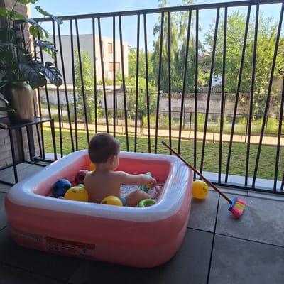 On a balcony in Mechelen, Belgium, a young child plays in a pink inflatable pool filled with water and several colorful balls. The child is facing away from the camera, reaching for a yellow toy. A broom with pink and yellow bristles leans against the pool. The balcony is tiled and overlooks a grassy area with trees and a building in the background. A potted plant with large leaves is visible to the left of the scene.