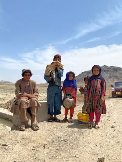 Five individuals, likely children, are located in a rural setting with sparse vegetation, possibly near Ashoqah, Afghanistan. An older male sits on a concrete structure adjacent to a water channel, wearing traditional clothing and sandals. Another male stands with a scarf covering part of his face. A girl in a blue headscarf holds a metal container. Another child stands slightly behind them. A girl in a floral patterned garment and headscarf stands to the right. A yellow bucket labeled "GREASE CITGO" is on the ground near the children. A vehicle is partially visible in the background, near a hill. The sky is blue with scattered clouds. The ground appears dry and dusty.