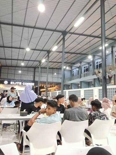 The image displays a lively indoor dining establishment or food court, identified as being in Kampongbaro, Indonesia. The venue features a high, vaulted corrugated metal roof supported by dark grey structural beams and columns, illuminated by numerous overhead circular and linear light fixtures. To the left, a prominent dark sign with white text reads "ELITE COFFEE." To the right, a multi-story building section with balconies and light grey walls featuring a white lattice pattern is visible, with several wall-mounted fans affixed to the columns.

The scene is occupied by a diverse group of individuals, predominantly young adults, seated at white rectangular tables on white plastic chairs. In the foreground, a young male with short dark hair, wearing a light blue polo shirt, sits at a table, looking towards the left of the frame. His right arm is bent at the elbow, resting on the table, with a watch visible on his wrist. A small black rectangular object rests on the table in front of him. To his right, a male in a grey long-sleeved shirt and another male in a dark patterned shirt are seated with their backs to the camera. In the mid-ground, a young woman wearing a light blue hijab and a denim jacket is seated, looking towards the left. Behind her, a woman in a light purple hijab stands with her back to the camera, near a male wearing a light green t-shirt and a dark cap. Numerous other patrons, many wearing head coverings, are dispersed throughout the background, seated at tables, engaged in dining and conversation. Various drinks and food items are visible on the tables.