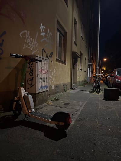In Duisburg, Germany, at night, a white and green electric scooter is parked on a sidewalk next to a beige building with graffiti. The building's wall is covered in various graffiti tags. To the right of the scooter is a grey electrical box, also covered in graffiti. The sidewalk is paved with concrete slabs and a metal grate is visible. Further down the sidewalk, a row of parked cars lines the street, including a red car. Several bicycles are parked near a lamppost. The scene is illuminated by a nearby streetlight and the light from the building.