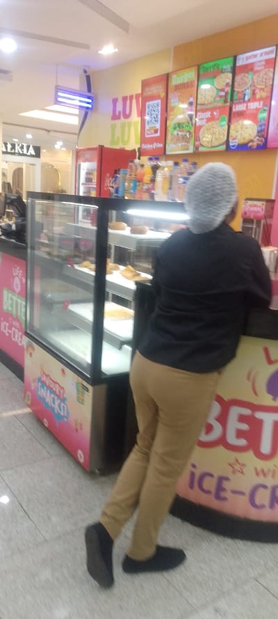This image depicts an indoor food service establishment located within a larger commercial complex, possibly a shopping mall, in Majengo ward, Kenya.

A sole individual, viewed from behind, is leaning against a food service counter in the foreground. This person is wearing a black long-sleeved shirt, tan/khaki trousers, black flat shoes, and a white hairnet completely covering their head. Their left foot is slightly lifted off the floor, resting on the toe.

The counter features a glass display case on the left, containing multiple shelves with various golden-brown pastries and round, bread-like items. The side panel of this counter displays branding, including "BETTER with ICE-CREAM" and "SAVOURY SNACKS!". Behind the individual, on the counter surface, are rows of assorted bottled beverages, including water, juices, and soft drinks. A red Coca-Cola branded refrigerator is partially visible to the left of the bottled drinks.

The background consists of a brightly lit wall with multiple illuminated menu boards. Prominent branding includes "LUV LUV" in large, colorful letters. The menu boards display items such as "Burger BOMBA!", various pizzas ("Classic Deal," "Large Triple"), and QR codes associated with "Chicken Inn." A blue-glowing insect zapper or light fixture is suspended from the ceiling. Further in the background, to the left, is the entrance to a store labeled "GALERIA," with display mannequins visible inside. The floor is covered with light-grey tiles.
