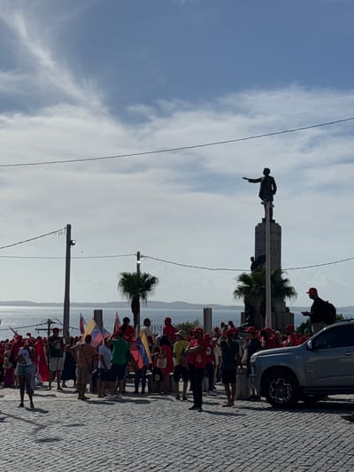 A statue stands atop a stone pedestal against a cloudy sky. A crowd, many wearing red hats, gathers along a cobblestone path near the water. Some hold Venezuelan flags. A parked silver vehicle is visible. Utility poles and palm trees line the path.