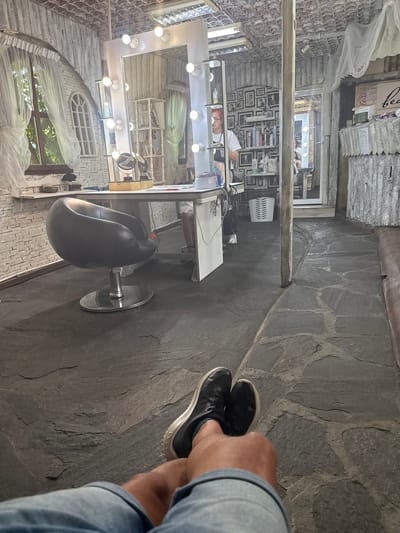The image depicts the interior of what appears to be a hair salon or beauty parlor. The flooring is made of irregular, large, dark grey stones, with some lighter grey grout visible between them. The walls are clad in a textured, light-colored material, possibly wood or concrete, with a corrugated appearance. The ceiling is also textured and light-colored, with visible light fixtures.
In the foreground, a person's legs and feet are visible, clad in denim shorts and black sneakers. The person is seated, with their legs crossed.
The main area of the salon features a large mirror with a vanity lighting setup, surrounded by decorative brickwork. A sleek, black salon chair with a chrome base is positioned in front of a white desk. On the desk, there are various items, including a wooden box, a mirror, and some bottles. Behind the mirror, shelves are stocked with hair products and decorative items, including framed pictures on the wall.
A man with red hair and glasses, wearing a white t-shirt and an apron, is visible in the background, seemingly working at the vanity. To the right of the vanity area, a doorway leads to another room, with more shelving and framed pictures visible. Further to the right, a portion of a tan leather sofa is visible.
The environment appears to be a rustic or bohemian-style salon, possibly located in Peshtera, Bulgaria, given the contextual information. The lighting is bright and functional, with multiple bulbs around the mirror. The overall impression is of a well-equipped and aesthetically distinct workspace.