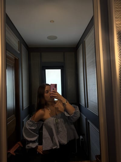 A young woman is taking a mirror selfie in a hallway. She is wearing an off-the-shoulder blue and white striped blouse and black pants. She has long brown hair and is wearing a necklace and a bracelet. She is holding a pink phone up to take the picture. The hallway has dark wood doors and patterned wallpaper. There is a toilet paper dispenser visible in the background. The location is identified as Santiago Yancuitlalpan, Mexico.