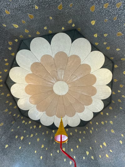 This is a low-angle, full-shot photograph taken from inside a circular or octagonal room in Chang Khoeng, Thailand. The ceiling is elaborately decorated with a large, multi-layered floral design in shades of cream and light brown. The floral pattern is composed of numerous petal shapes radiating from a central cream-colored circle. The walls and the portions of the ceiling not covered by the floral design are dark gray and speckled, with numerous gold leaf cutouts of small leaves scattered across the surface. A yellow conical lantern with a red pole hangs from the ceiling, positioned towards the bottom right of the frame. No people are visible in the image. The overall impression is one of intricate and ornate architectural decoration.
