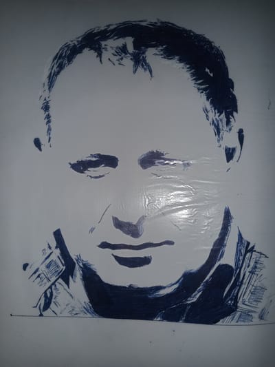 This is a blue ink drawing of a man's face on a white, textured background. The drawing is somewhat abstract and appears to be done in a sketchy style, with bold lines and shading used to create the features. The man's eyes are closed or heavily lidded, and his mouth is slightly downturned. He has short, dark hair. The drawing is cropped to show primarily the face and the upper chest area. The background shows some texture that might suggest a surface like a woven fabric or a textured paper. The image is lit from the right, creating a slight glare on the surface of the drawing, particularly across the man's forehead and right cheek. There are no discernible elements in the image that indicate a specific location in the city of Gulbag, Uzbekistan, or any other urban environment.