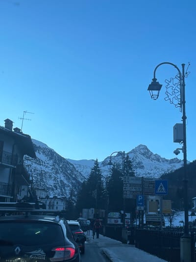 The image captures a vibrant street scene in La Thuile, Italy, set against a stunning backdrop of snow-capped mountains under a clear, bright blue sky. The scene depicts a sunny, cold day, likely late morning or early afternoon, given the clear illumination.

In the foreground, a prominent street light with decorative snowflake-like metalwork extends over the road. A surveillance camera is attached to the light pole. Below, several vehicles are present on the street, including a dark Renault car in the lower right, which has a "LA THUILE SNOWBIKE" sticker on its rear window. Buildings with alpine architectural styles line the road, interspersed with dark evergreen trees that also dot the lower slopes of the mountains.

On the right side of the street, a cluster of informational and directional signs is visible. These signs include a pedestrian crossing symbol, a parking symbol ("P"), and text referring to locations such as "FAUDOUZE," "Cafe Tableau," and "Il Mulino," along with various directional arrows. Faint figures of pedestrians can be seen in the distance near these signs. The snow-covered landscape and clear weather indicate a typical winter day in this Italian mountain resort.