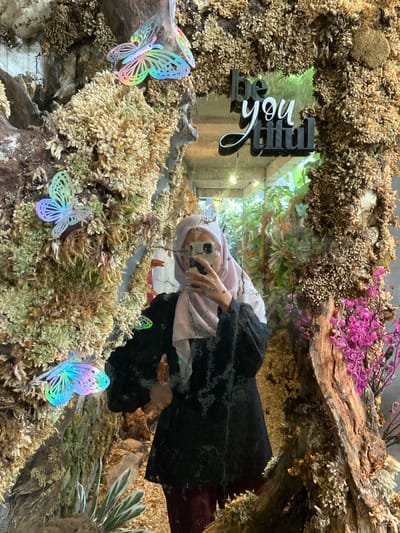 A young woman with light skin, wearing a light pink hijab and a black dress, is taking a selfie in a mirror. The mirror is framed by a rustic, textured surface resembling bark and moss, adorned with iridescent butterfly decorations. The background reflected in the mirror shows a lush indoor plant display and the words "be you tiful" in raised lettering. The scene suggests an indoor, possibly decorative, setting like a cafe or shop. The location is identified as Rengat, Indonesia.