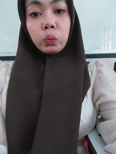 This is a close-up, vertically oriented selfie of a young woman wearing a dark brown hijab. She is wearing makeup, including mascara and lipstick. Her head is tilted, and she is looking at the camera with her lips slightly parted. Behind her, a light-colored garment with embroidered green leaves and a small white flower is visible. The background appears to be an indoor setting with a white wall or whiteboard and some windows with horizontal blinds. The lighting suggests it could be daytime. The woman's expression is neutral. There is no visible text in the image. The scene is likely a personal moment captured by the individual.