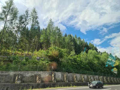 This image captures a view from a moving vehicle on a highway, likely in or near St. Barbara im Mürztal, Austria, given the surrounding forested hills. The scene is dominated by a dense forest of evergreen trees ascending a steep slope to the left and center. A grey concrete barrier, designed to prevent rockfalls or debris from reaching the road, runs horizontally across the lower portion of the image. This barrier is composed of large, pre-cast concrete blocks, many of which have integrated square indentations or openings. Above this barrier, a chain-link fence separates the road from the forested embankment.
In the foreground, a grey hatchback car is traveling on the highway. The sky is a mix of bright blue patches and white, fluffy clouds. The overall impression is a typical roadside view of a mountainous, wooded region.