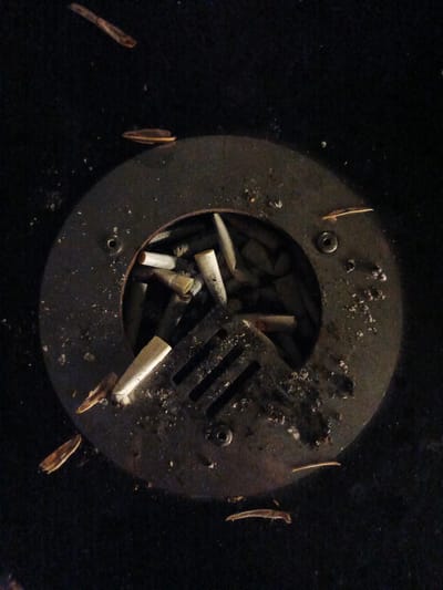 The image shows a close-up, overhead view of a metal ashtray filled with cigarette butts. The ashtray is circular with a grate in the center and is set into a dark, textured surface that appears to be outdoor carpeting or a similar material. Small pieces of debris are scattered around the ashtray. Several long, thin, brown objects are scattered on the dark surface surrounding the ashtray, some appearing to be leaves or twigs. The lighting is dim, casting shadows and highlighting the texture of the objects. The ashtray and its contents suggest an urban or public outdoor space. There are no people or animals visible in the image. The location is specified as Sof'ino, Russia.