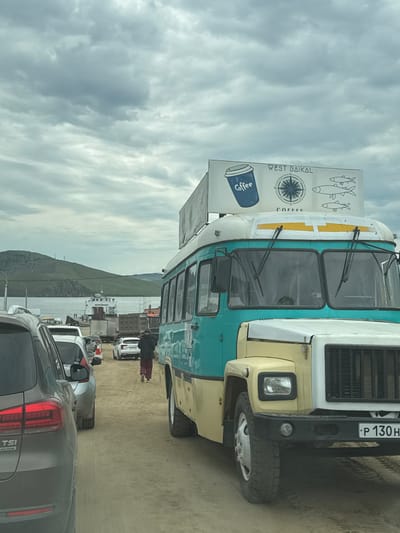 The image captures a view of a ferry waiting area at Lake Baikal, near the city of Khuzhir, Russia. The foreground is dominated by a custom-built vehicle, appearing to be a converted bus or truck, painted teal and cream, with a sign on top depicting a coffee cup, a compass, and fish, advertising "WEST BAIKAL COFFEE." A line of cars, including a grey SUV in the immediate foreground, are waiting on a sandy surface. In the mid-ground, a ferry, identified by the Cyrillic text "ДОРОЖНИК" (Dorozhnik), is docked. The background features a body of water, likely Lake Baikal, and rolling green hills under a cloudy sky. A single individual, dressed in dark clothing and maroon pants, walks away from the camera towards the ferry. The overall scene suggests a moment of transit and commerce at a ferry terminal.