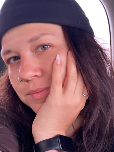 A close-up, selfie-style portrait shows a person wearing a black beanie and a dark top. The person has light skin with freckles, green eyes, and brown hair. Their left hand is raised to their face, with a manicured fingernail visible. They are wearing a silver nose ring and a silver ring on their right ring finger. A black smart watch is visible on their left wrist. The background is blurred, suggesting an indoor or vehicle setting with a bright light source on the right. The image does not contain any contextual information regarding Petrovskoye, Russia.
