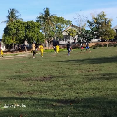 The scene is a grassy field in Nungwi, Tanzania, at coordinates -5.726 latitude and 39.301 longitude. Seven people are playing soccer. Five of them are wearing yellow shirts and black shorts, with two wearing black shirts and blue shorts. The players are on a field surrounded by trees and a two-story building with a white roof. The sky is clear blue. The person taking the picture is standing in the grassy field with a low angle. The image shows a typical day in Nungwi with people enjoying outdoor activities. The field is bordered by trees and a white building with a white fence. The image was taken with a Galaxy A05s phone. The field is grassy and brown in some spots.